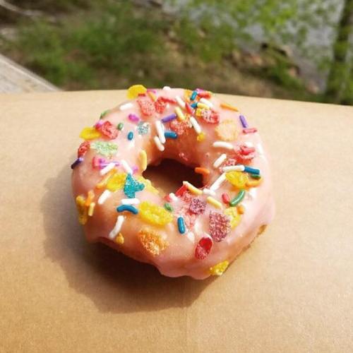 A pink frosted donut topped with colorful sprinkles, resting on a brown paper surface with greenery in the background.