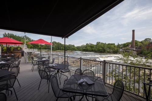 Outdoor dining area with black tables and red umbrellas overlooking a river and waterfall. Lush greenery in the background.