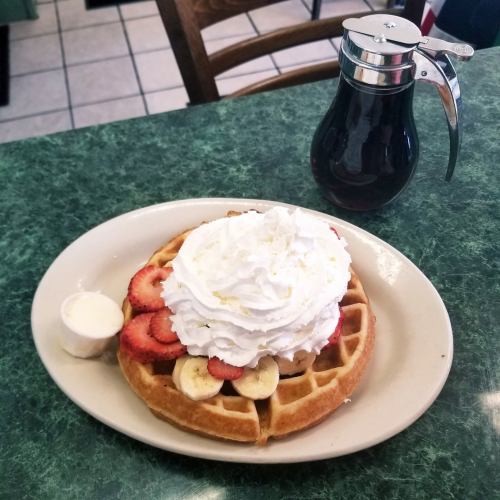 A waffle topped with strawberries, bananas, and whipped cream, served with a side of syrup and a small cup of cream.