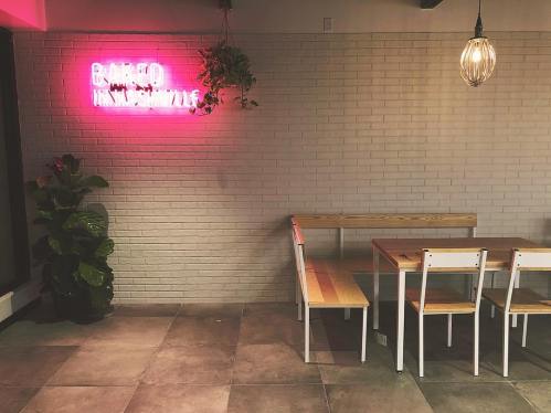 A cozy cafe interior with a pink neon sign, wooden benches, and a potted plant against a brick wall.