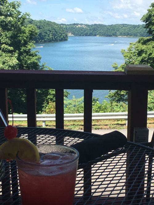 A refreshing cocktail sits on a table with a scenic view of a lake and lush greenery in the background.