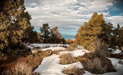 A scenic landscape featuring snow-covered ground, rocky terrain, and evergreen trees under a partly cloudy sky.