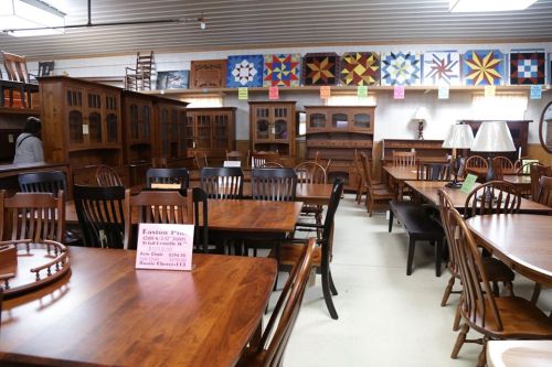 A furniture store interior featuring wooden tables, chairs, and cabinets, with colorful quilts displayed on the wall.