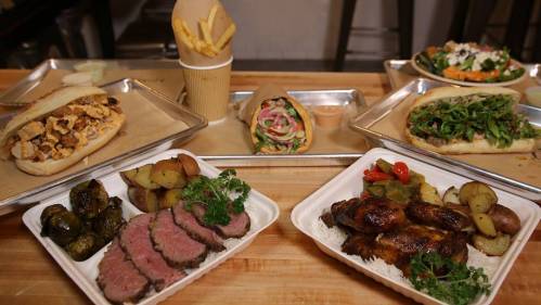 A variety of dishes on metal trays, including sandwiches, fries, roasted vegetables, and grilled meats.