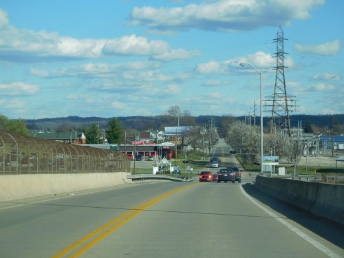 A view of a wide road with cars, power lines, and distant hills under a blue sky with fluffy clouds.
