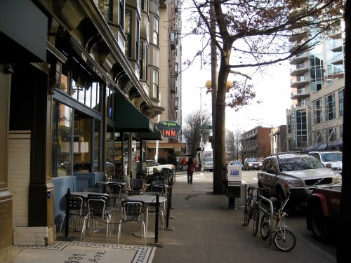 A city street scene featuring a café with outdoor seating, parked bicycles, and nearby buildings.