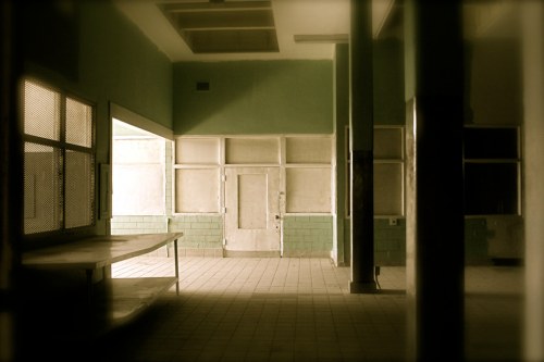 Abandoned interior of a room with green walls, large windows, and empty tables, creating a desolate atmosphere.