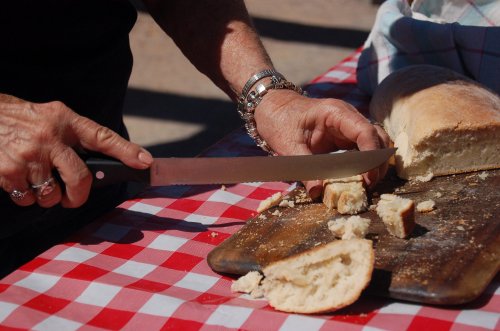 A person slices a loaf of bread on a wooden cutting board, with pieces scattered on a red and white checkered tablecloth.