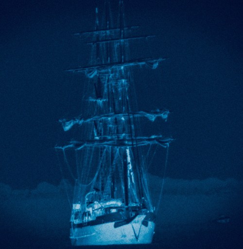 A tall ship illuminated in blue light, with multiple masts and sails, set against a dark, cloudy background.