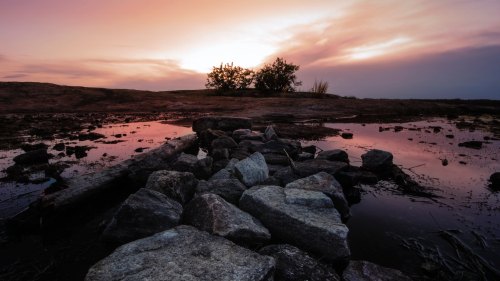 A serene landscape at sunset, featuring a rocky path leading to a calm water body and silhouetted trees in the background.