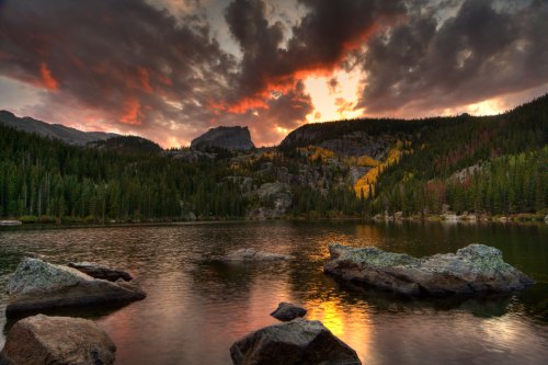 A serene lake surrounded by mountains and trees, reflecting a colorful sunset with dramatic clouds overhead.