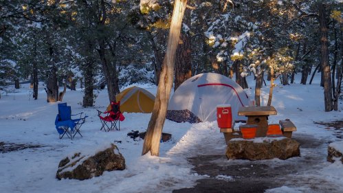 A snowy campsite with two tents, camping chairs, a picnic table, and a cooler surrounded by trees.