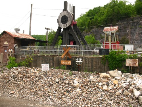 A vintage mining structure with a large wheel, surrounded by old signs and a rocky area, set against a green hillside.