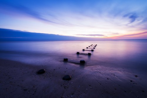 A serene beach scene at sunset, featuring a row of wooden posts leading into calm waters under a colorful sky.