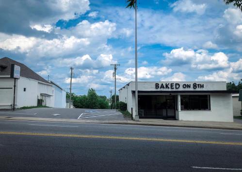 A white building with "BAKED ON 8TH" sign, set against a cloudy sky and empty parking lot.