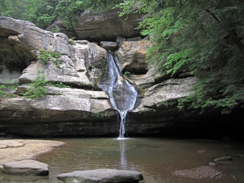 A serene waterfall cascading over rocky cliffs into a calm pool, surrounded by lush greenery and towering trees.
