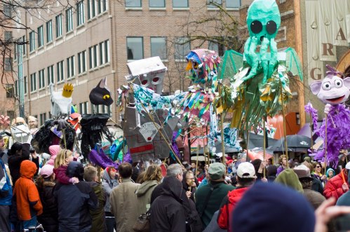 A lively parade scene with colorful puppets and a crowd of people in winter clothing enjoying the festivities.