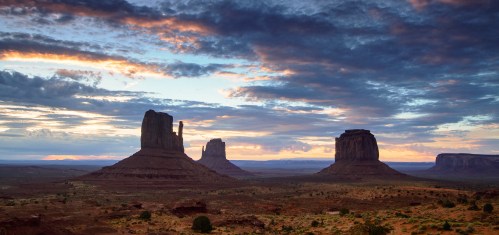 A stunning desert landscape at sunset featuring iconic rock formations under a colorful sky with scattered clouds.