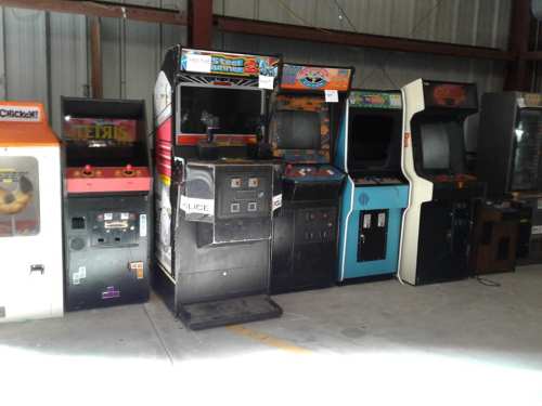 A row of vintage arcade machines in a warehouse, including games like Tetris and Steel Runner.