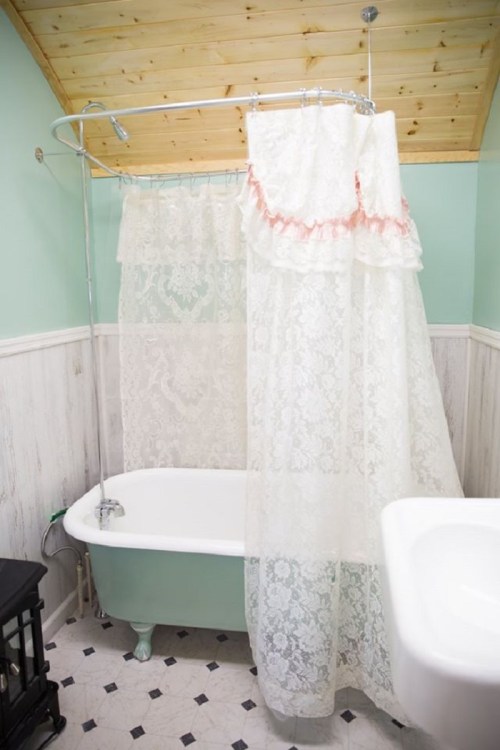 A vintage bathroom featuring a clawfoot tub, lace shower curtain, and light green walls with wooden accents.