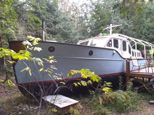A vintage boat resting on land, surrounded by lush greenery and trees, with a wooden deck nearby.