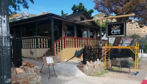 A quaint café with a wooden porch, surrounded by trees and stone walls, featuring outdoor seating and construction scaffolding.