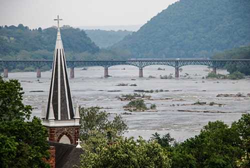 A church steeple in the foreground with a river and bridge surrounded by green hills in the background.