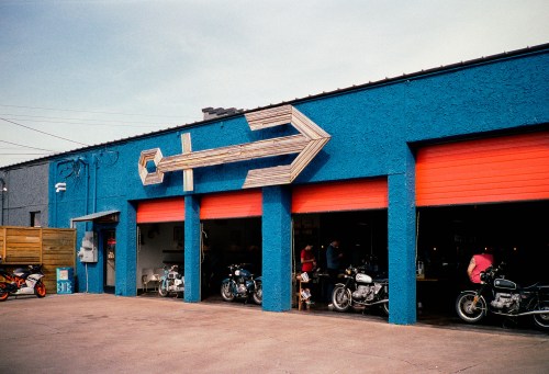 A vibrant blue building with orange garage doors and a large wooden arrow sign, featuring several motorcycles outside.