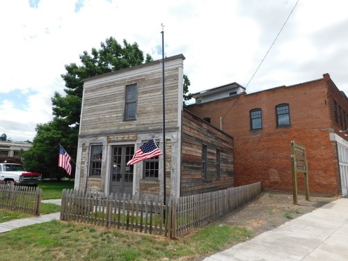 Historic wooden building with American flags, surrounded by a picket fence and greenery, set against a cloudy sky.