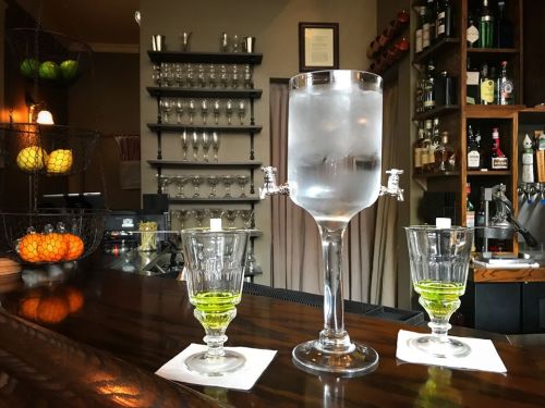 A bar scene featuring a large glass dispenser filled with ice water, flanked by two small glasses of green liquid.