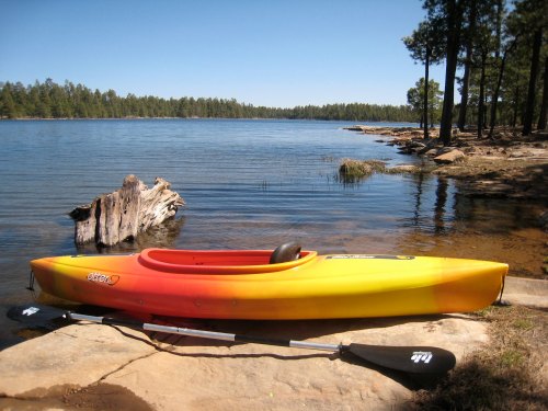 A bright orange and yellow kayak rests on a rocky shore by a calm lake, surrounded by trees and clear blue skies.
