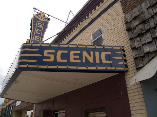 Sign for the Scenic theater, featuring bold blue and gold lettering, mounted on a brick building.