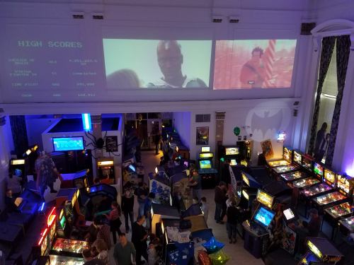 A vibrant arcade filled with pinball machines and classic games, featuring high scores displayed on the wall and movie projections.