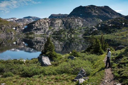 A person stands on a trail by a serene lake, surrounded by mountains and lush greenery under a clear blue sky.