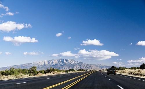 A wide highway stretches towards distant mountains under a blue sky with fluffy clouds.