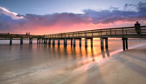 A serene sunset over a pier, with vibrant colors reflecting on calm waters and gentle waves lapping at the shore.