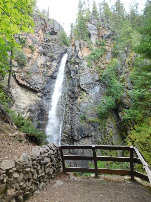 A tall waterfall cascades down rocky cliffs, surrounded by lush green trees and a wooden railing in the foreground.