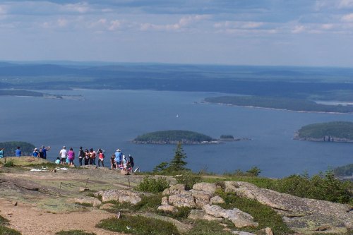 A group of people stands on a rocky overlook, gazing at a scenic view of islands and water under a blue sky.