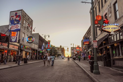 A lively street scene at sunset, featuring neon signs, shops, and people walking along a bustling urban area.