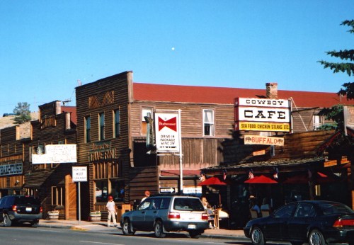 A street view of rustic buildings, including a café and shops, under a clear blue sky. Cars are parked nearby.