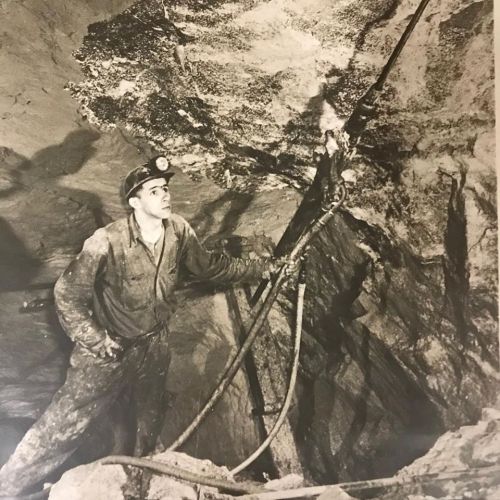 A miner in a dark tunnel, wearing a helmet with a light, holding equipment and looking up at the rock formations above.