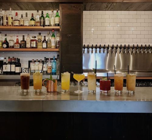 A bar counter displaying a variety of colorful cocktails and drinks, with shelves of liquor in the background.
