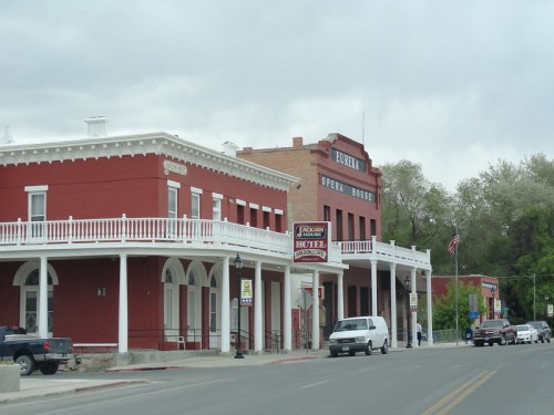 Historic buildings line a street, featuring a red hotel and an opera house under a cloudy sky.