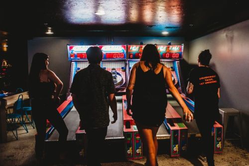 Four people stand in front of arcade skee-ball machines, preparing to play in a dimly lit game room.