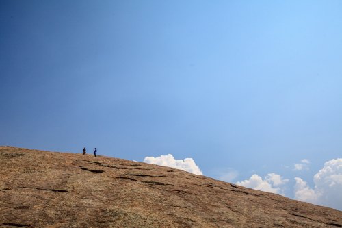 Two hikers stand on a large, rocky surface under a clear blue sky with a few clouds in the distance.