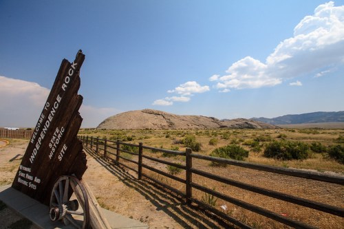 Sign for Independence Rock with a rocky formation in the background and a wooden fence in a grassy landscape.
