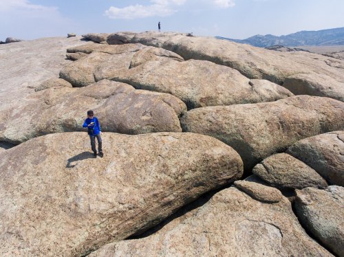 Two people stand on large, smooth boulders under a blue sky, with distant mountains visible in the background.