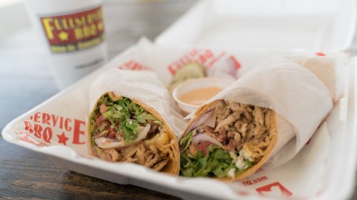 Two wrapped burritos filled with shredded meat, lettuce, and tomatoes, served in a takeout container with a dipping sauce.