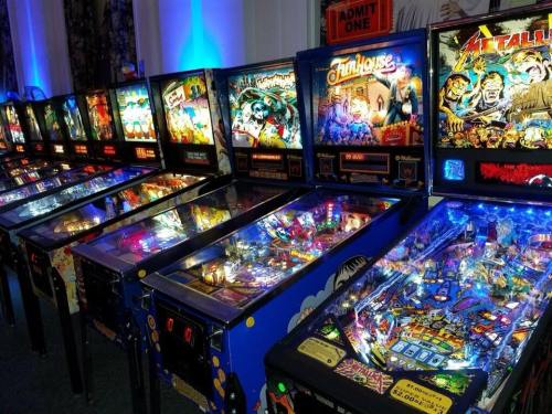 A row of colorful pinball machines, each with unique designs and illuminated playfields, in a vibrant arcade setting.