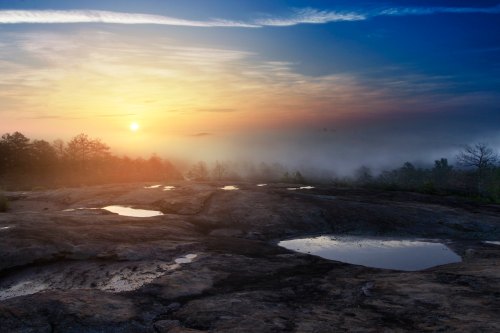 A serene sunrise over a misty landscape, with reflections in puddles on rocky terrain and trees in the background.